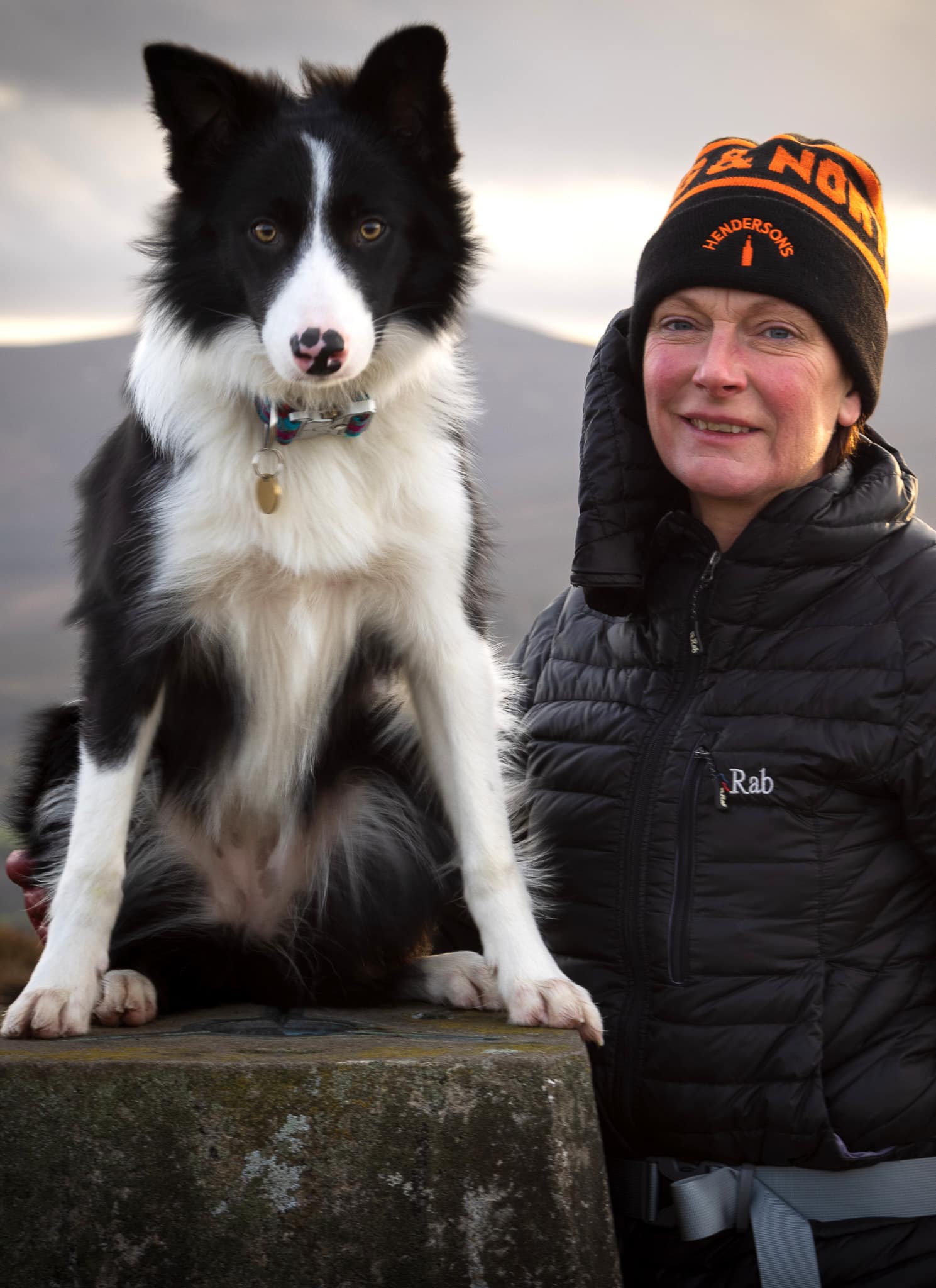 A portrait photograph of a lady and her border collie dog standing together on top of a hillside in Aberdeenshire, capturing their joyful expression against a stunning natural backdrop, taken by Julian Davies Pet Photography