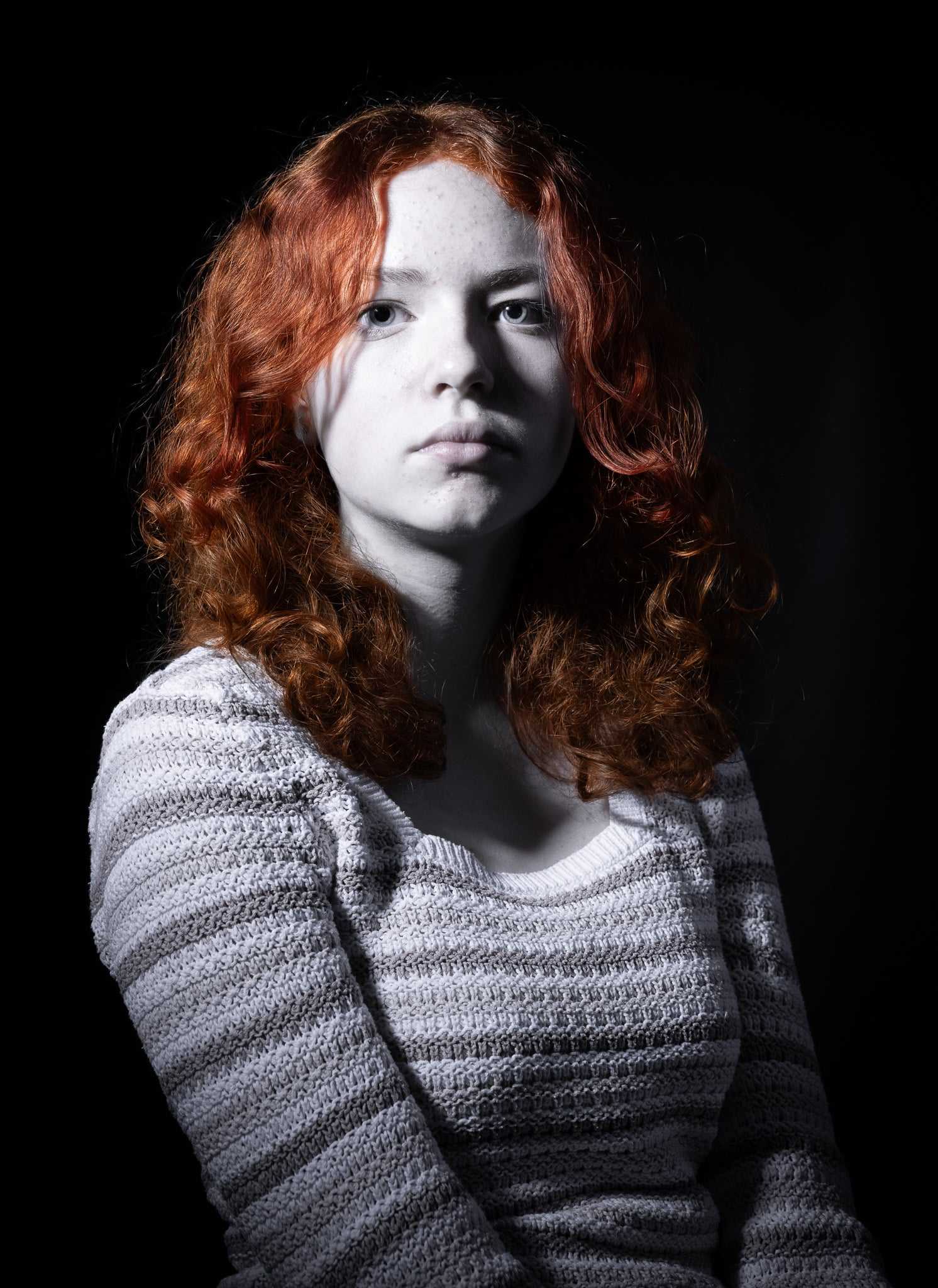 A black and white photograph of a young lady with a sullen expression, gazing directly at the camera, with only her hair highlighted against a dark background, captured in the Julian Davies Photography studio based in Aberdeenshire.